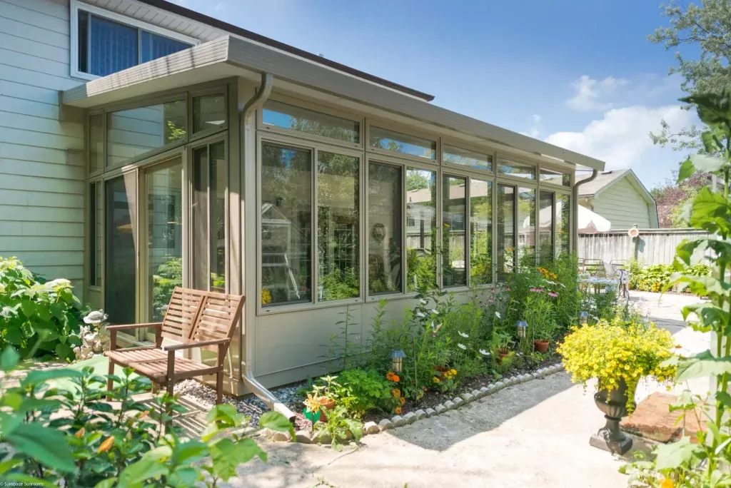 Tan 3 season glass sunroom addition attached to a house with a garden walkway