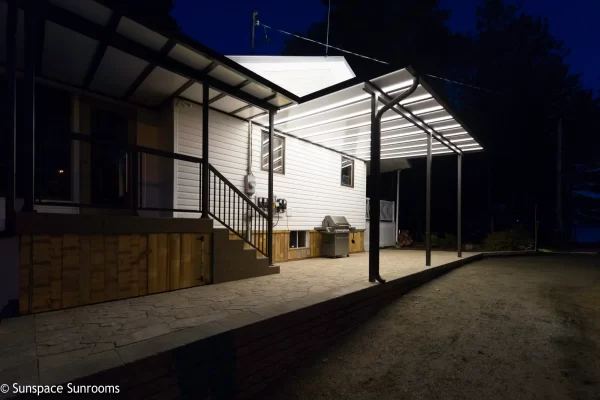 Illuminated patio cover with dark metal framing installed over stone paver outdoor deck at night